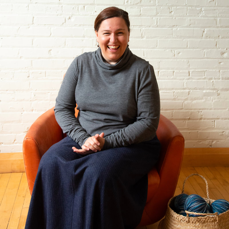 LISA AVIVA's knitter, Erica, is pictured seated and against a painted white brick wall. Erica is wearing LISA AVIVA's Tailored Turtleneck, in Slate and the Full Wool Skirt, in Night Pinstripe.